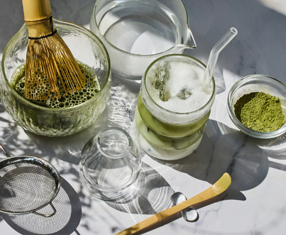  A layered iced ceremonial matcha latte on a sunlit table next to organic green tea powder and a bamboo whisk, representing a clean energy ritual.