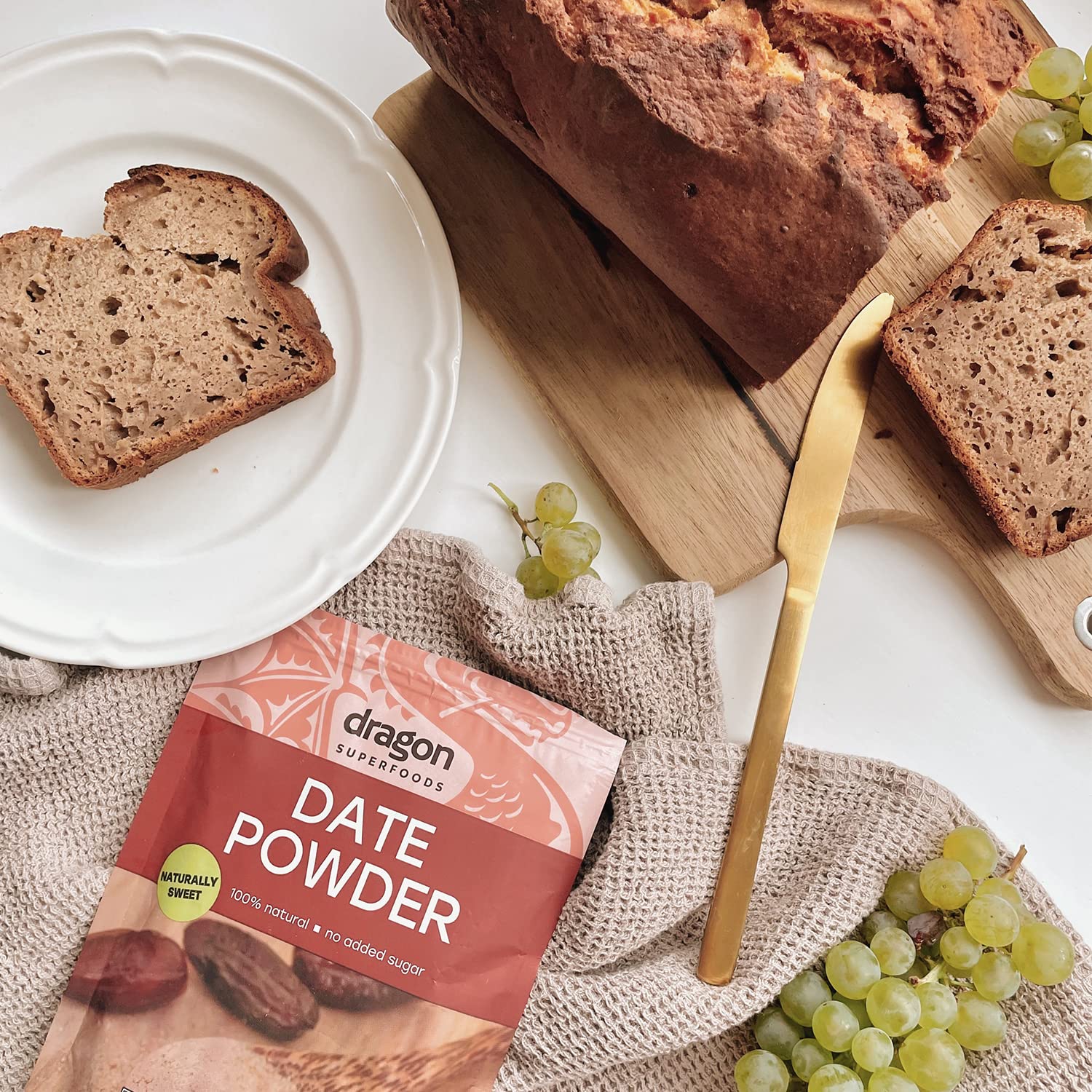 Loaf of bread with slices, grapes, and a package of Dragon Superfoods Date Powder on a white surface.