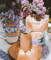Maca powder with a glass of a pinkish drink and flowers on a table.